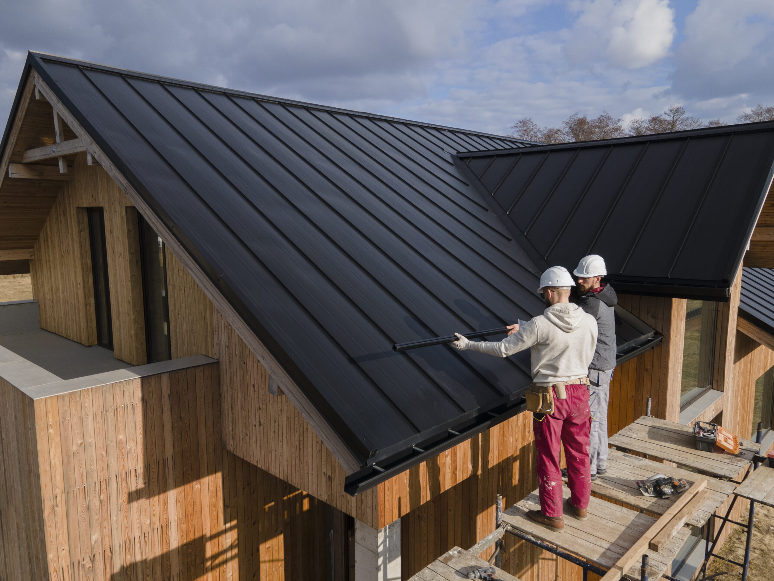 Construction workers in hard hats working together to install a metal roof on a wooden house, showcasing teamwork and craftsmanship in building the house.