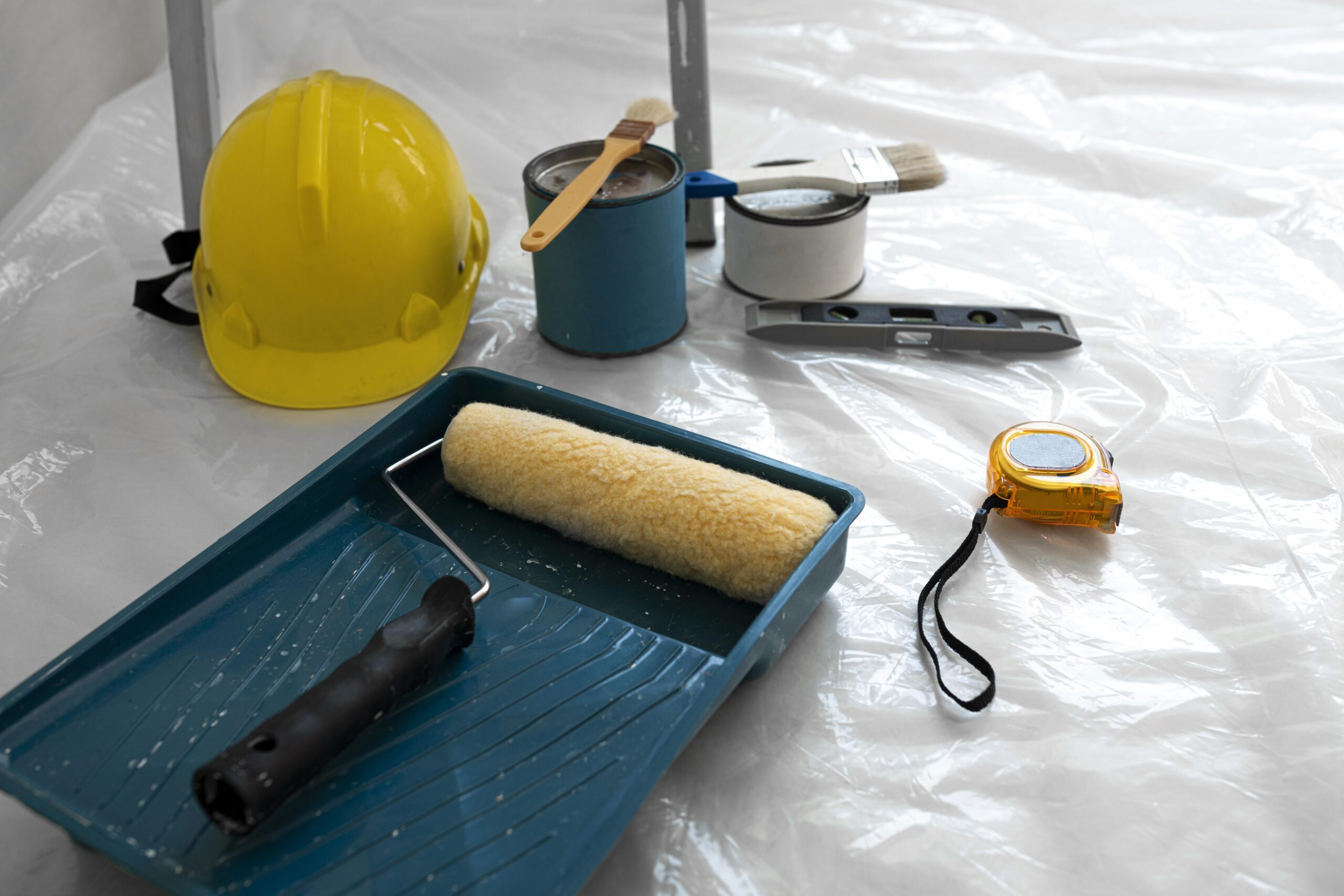 Construction tools including a yellow hard hat, paint rollers, brushes, and a tape measure on a plastic sheet, representing the tools used in Pre-Construction Services.