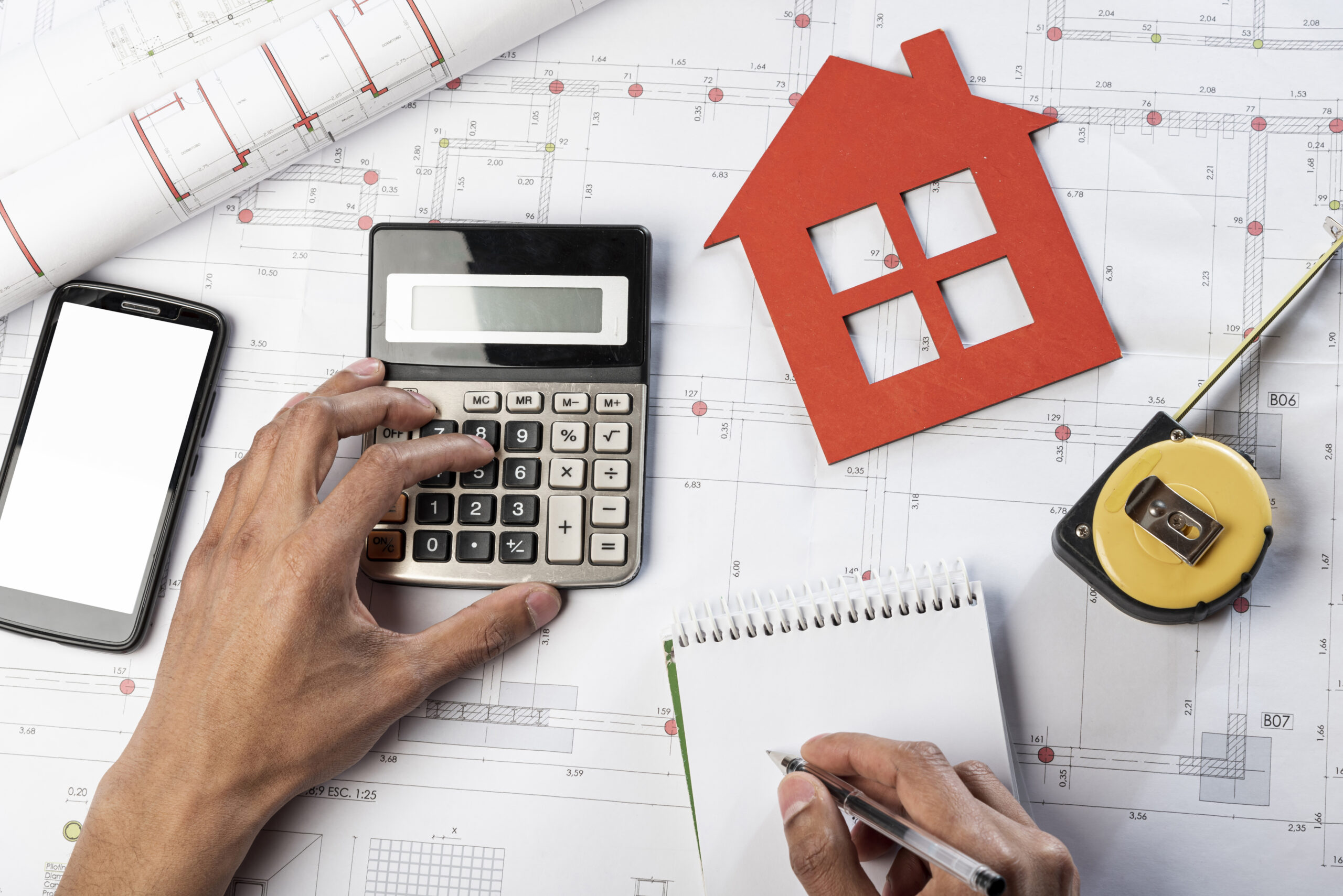 A person is using a calculator and writing notes on a notepad while reviewing construction blueprints, with a red house cutout and measuring tape placed on the table.