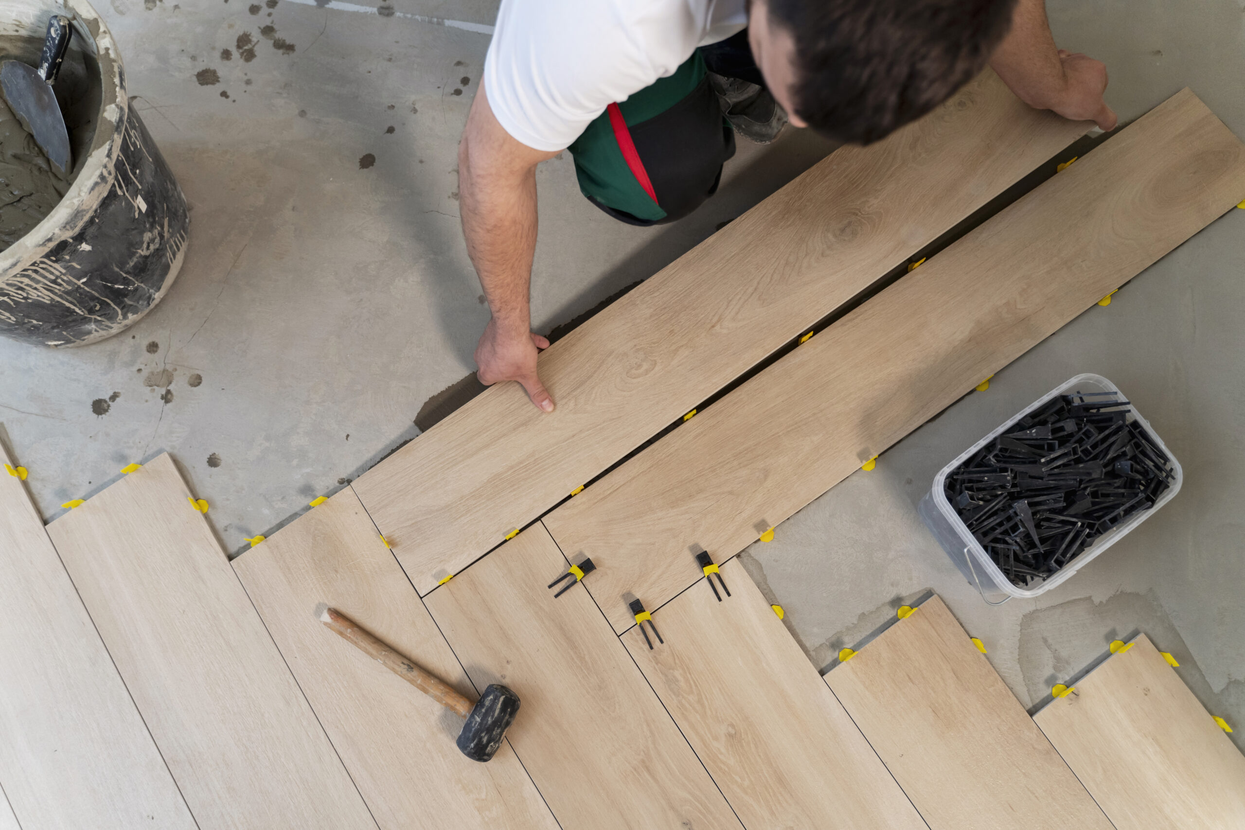 A person is laying wooden floor tiles on the floor, using a hammer and spacers to align the tiles, with a bucket of grout nearby