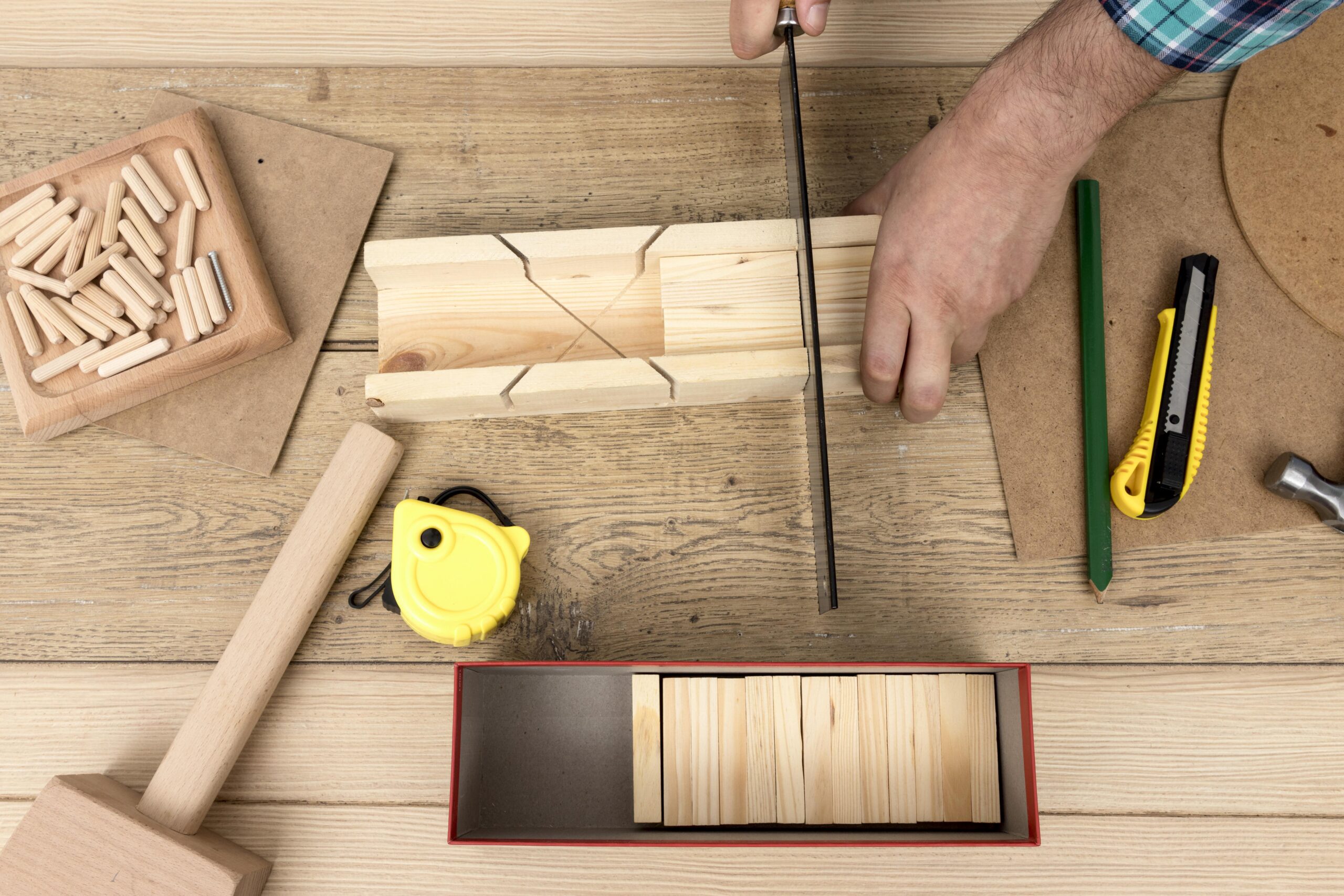 A person is measuring and cutting wood using a saw, with various woodworking tools such as a hammer, pencil, measuring tape, and wooden pegs on a wooden workbench.